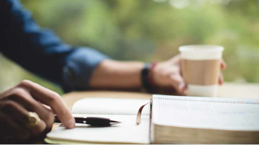 A person journaling about books, writing in a notebook with a cup of coffee.